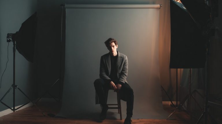 Man in a blazer sitting on a stool in front of a photography backdrop with studio lights on each side